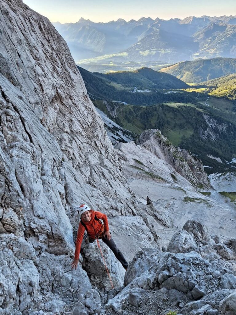 Ein Kletterer in orangefarbener Jacke und mit Helm erklimmt die felsige Wand des Pichlwegs am Dachstein, während sich unter ihm und in der Ferne eine weite Landschaft aus grünen Tälern und fernen Bergen erstreckt.