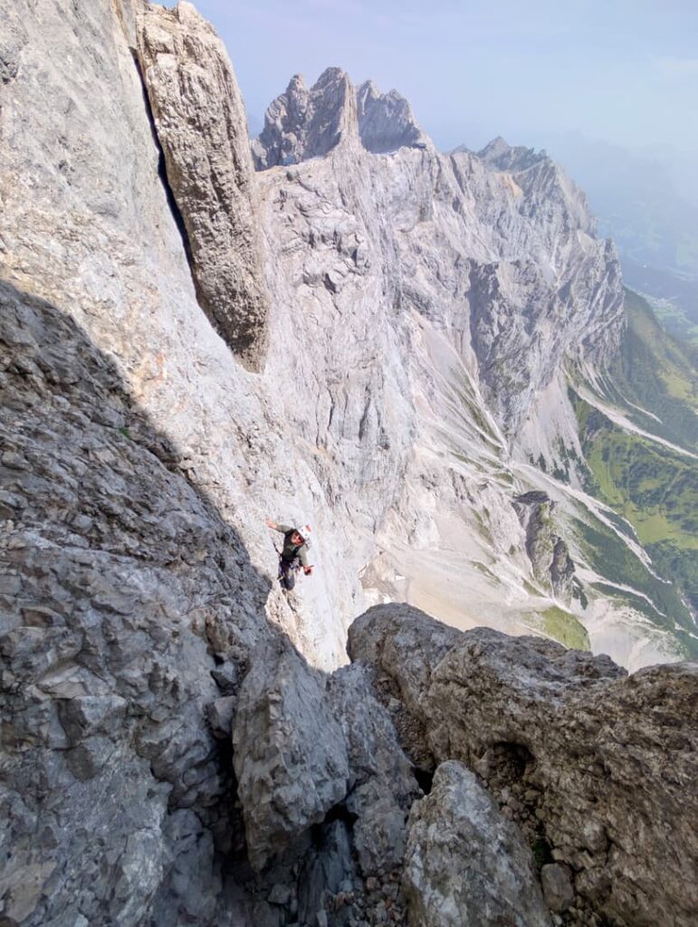 Ein Bergsteiger erklimmt einen steilen, felsigen Berghang entlang des berühmten Steinerwegs am Dachstein, während im Hintergrund unter einem dunstigen Himmel dramatische, zerklüftete Gipfel und ein entferntes Tal zu sehen sind.