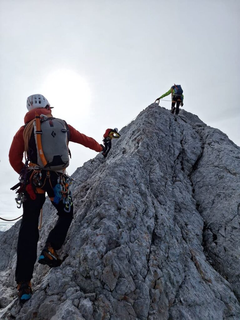 Drei Bergsteiger mit Helmen und Ausrüstung erklimmen unter einem strahlend bewölkten Himmel einen steilen, felsigen Berggrat; zur Sicherheit sind sie auf der berühmten Route „Steinerweg Dachstein geführt“ mit einem Seil miteinander verbunden.