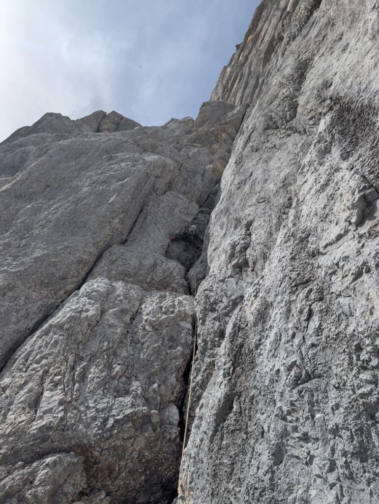 Ein Kletterer erklimmt unter teilweise bewölktem Himmel eine steile, graue, felsige Wand und bewältigt dabei die beeindruckende Route „Steinerweg Dachstein geführt“. Der Kletterer ist zwischen den großen Felsformationen teilweise zu sehen.