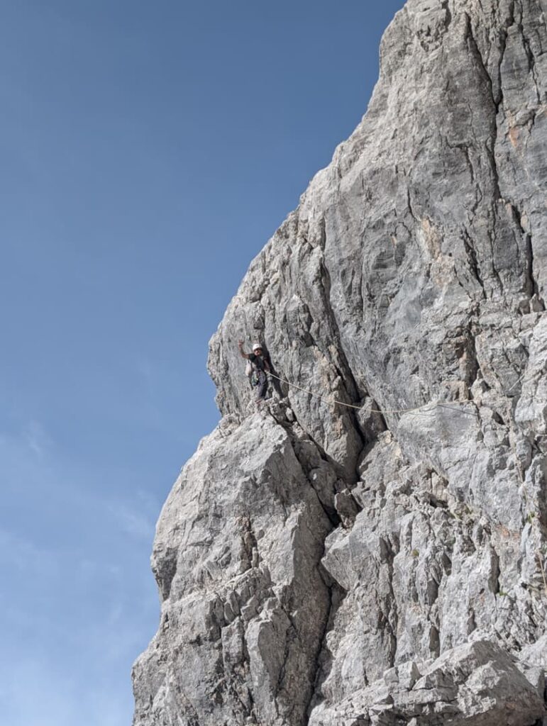 Ein Kletterer erklimmt unter strahlend blauem Himmel hoch über dem Boden eine steile, zerklüftete graue Felswand auf der Route „Steinerweg Dachstein geführt“.