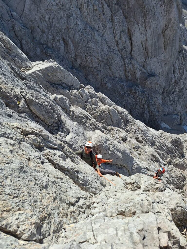 Drei Bergsteiger mit Helmen erklimmen eine steile, felsige Bergwand entlang der Route „Pichlweg Dachstein geführt“. Sie sind von schroffem Gelände umgeben, und das Sonnenlicht lässt die zerklüfteten grauen Felsen hervorstechen. Die Szene zeigt einen anspruchsvollen alpinen Aufstieg.