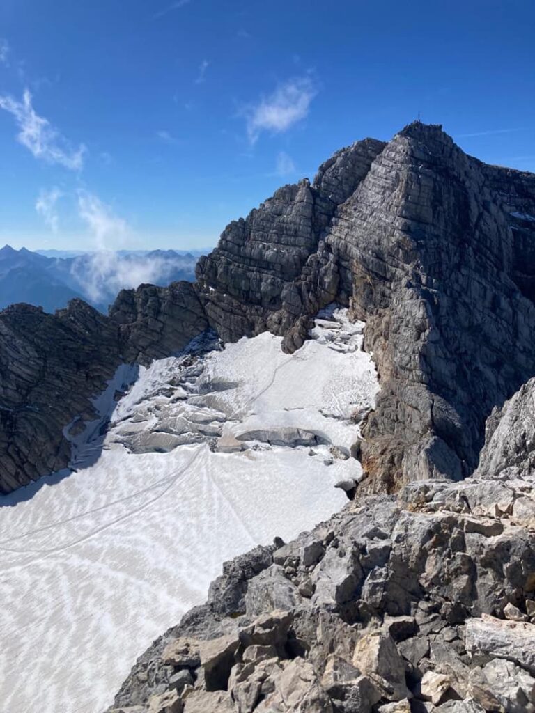 Ein Gipfel in den Rocky Mountains mit Schneeflecken unter strahlend blauem Himmel. Schroffe Felsen und Schneefelder bilden eine dramatische Landschaft, in der am Horizont entfernte Berge zu sehen sind – ein ikonischer Anblick, den oft diejenigen bewundern, die auf dem Steinerweg-Dachstein-Führungsweg durch diese Alpenregion wandern.