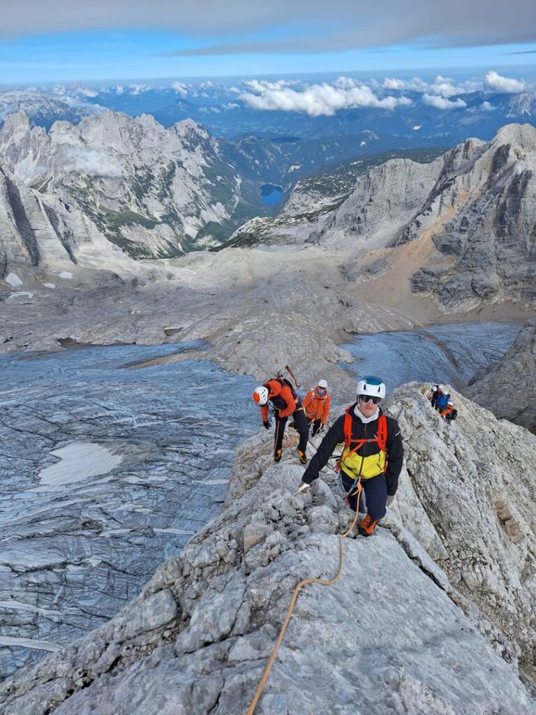 Vier Bergsteiger mit Helmen und Ausrüstung erklimmen einen felsigen Berggrat, aneinander geschnürt auf einer Route namens „Steinerweg Dachstein geführt“. Im Hintergrund sind unter einem teilweise bewölkten Himmel Schnee, schroffe Gipfel und ein weit entfernter See zu sehen.