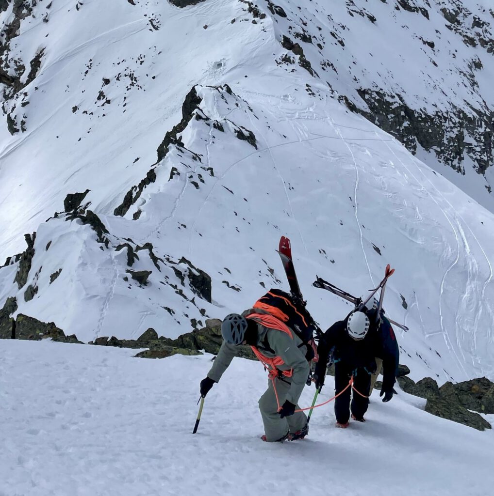 Zwei Bergsteiger in Winterausrüstung erklimmen mit Eispickeln einen steilen, verschneiten Berghang, wobei sie ihre Skier am Rucksack befestigt haben – ein Abenteuer, wie es oft auf geführten Bergtouren zu erleben ist. Im Hintergrund sind schneebedeckte Gipfel und Grate zu sehen.