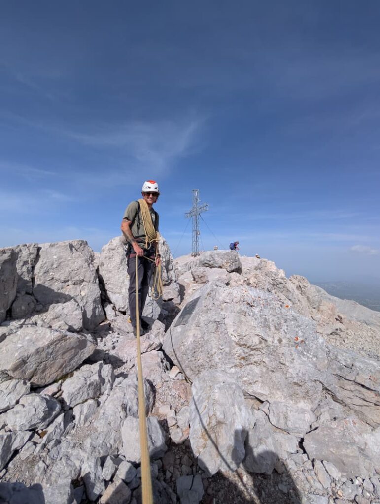 Ein Kletterer mit Helm steht auf felsigem Gelände und hält ein Seil in der Hand – eine Szene aus der „Steinerweg Dachstein geführt“-Besteigung. Im Hintergrund sind unter strahlend blauem Himmel eine weitere Person und ein großes Metallkreuz zu sehen.