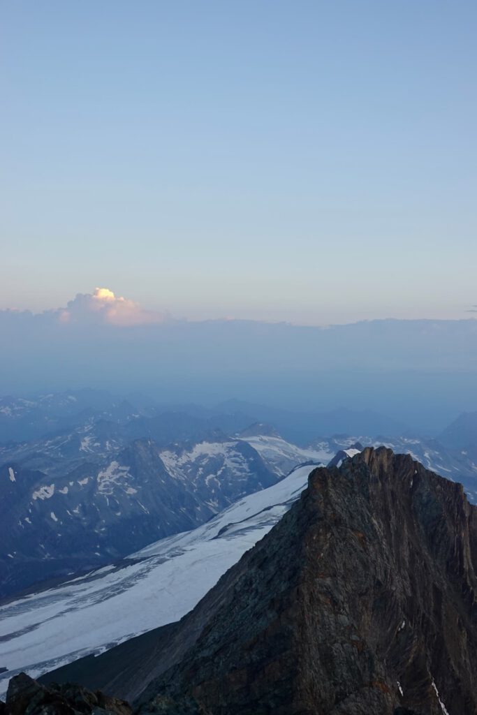 Ein felsiger Berggipfel im Vordergrund mit Schnee und Gletscher darunter, ferne Berge im Hintergrund und ein größtenteils klarer Himmel mit wenigen Wolken bei Sonnenaufgang oder Sonnenuntergang – diese atemberaubende Landschaft erinnert an die klassische Alpenkulisse entlang des Großglockner-Normalwegs.