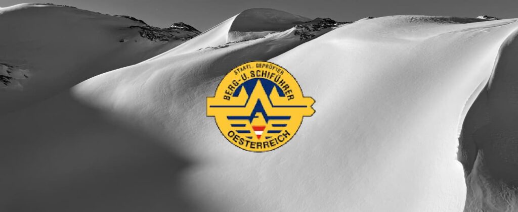 Ein gelb-blaues Abzeichen mit Schriftzug und einem Stern-Emblem, auf dem die Worte „staatlich geprüfte Bergführer“ zu lesen sind, ist in der Mitte einer schwarz-weißen verschneiten Berglandschaft abgebildet.