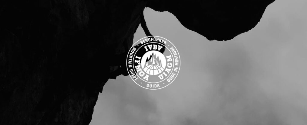 Die Silhouette eines Kletterers erklimmt eine steile Felswand; darüber ist vor einem bewölkten Himmel mittig ein rundes IFMGA-Logo für Bergführer zu sehen, auf dem der Schriftzug „staatlich geprüfte Bergführer“ hervorgehoben ist.