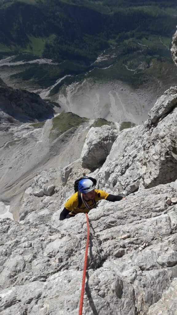 Ein Kletterer in gelbem T-Shirt und Helm erklimmt eine steile Felswand auf dem berühmten Kletterweg „Steinerweg Dachstein geführt“, gesichert durch ein rotes Seil, während weit unter ihm ein malerisches Tal und ein Wald zu sehen sind.