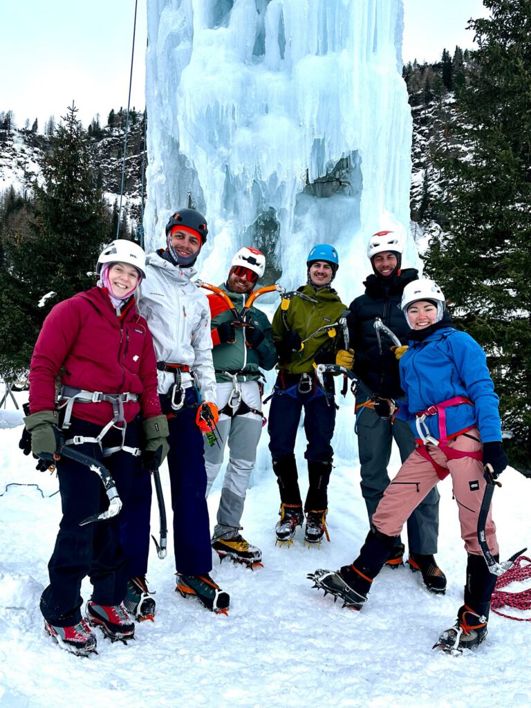 Sechs Personen in Winterkleidung und mit Helmen stehen lächelnd vor einem großen gefrorenen Wasserfall, umgeben von Schnee und Bäumen, und scheinen bereit für ihr Abenteuer im Eiskletterkurs Hohe Tauern zu sein.