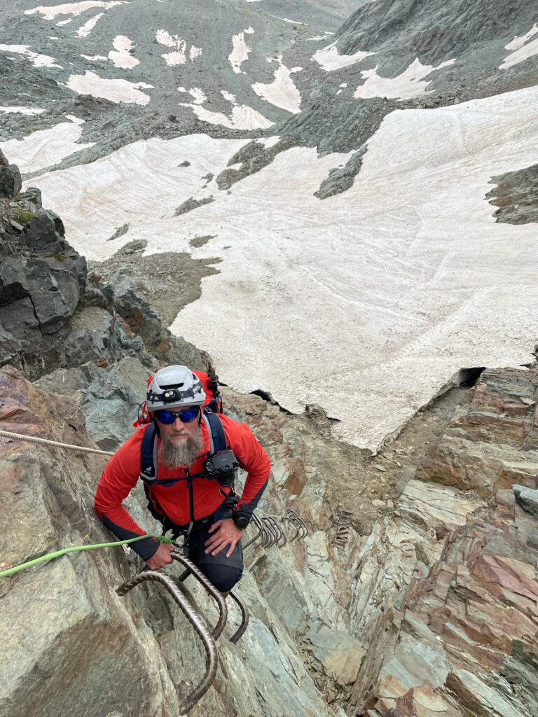 Ein bärtiger Bergsteiger in roter Jacke und mit Helm erklimmt auf dem „Großglockner Normalweg geführt“ mithilfe von Metallsprossen eine steile, felsige Bergwand, während unter ihm schneebedecktes Gelände und schroffe Gipfel zu sehen sind.