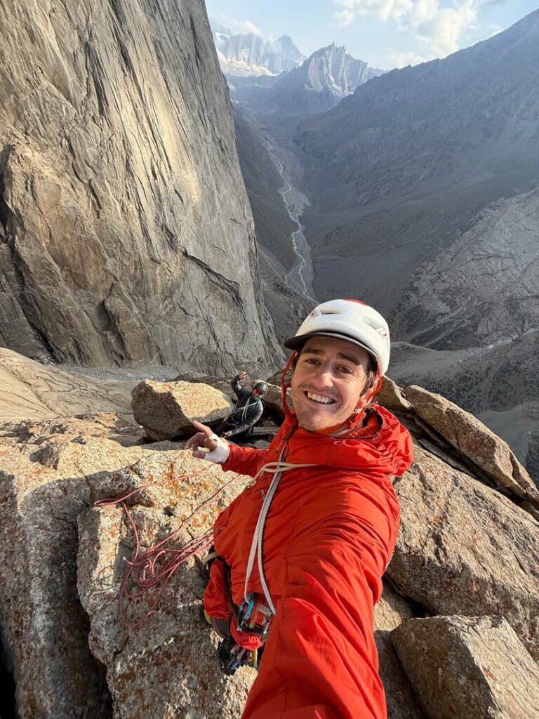 Ein Kletterer in roter Jacke und mit Helm macht auf einem felsigen Felsvorsprung lächelnd ein Selfie, hinter ihm steht ein weiterer Kletterer. Unter einem teilweise bewölkten Himmel erstrecken sich steile Klippen und ein tiefes Tal – eine atemberaubende Szenerie, die den Abenteuergeist einfängt, den Manuel Gratz Bergführer oft verkörpert.