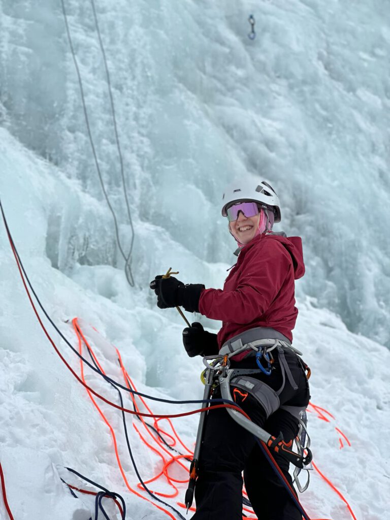 Eine Person in Winterkletterausrüstung steht auf Eis und lächelt, während sie im Rahmen eines Eiskletterkurses in den Hohen Tauern mit Seilen hantiert. Sie trägt eine rote Jacke, einen Helm und eine Schutzbrille, und an ihrem Klettergurt ist Kletterausrüstung befestigt. Auf dem verschneiten Boden liegen bunte Seile ausgebreitet.