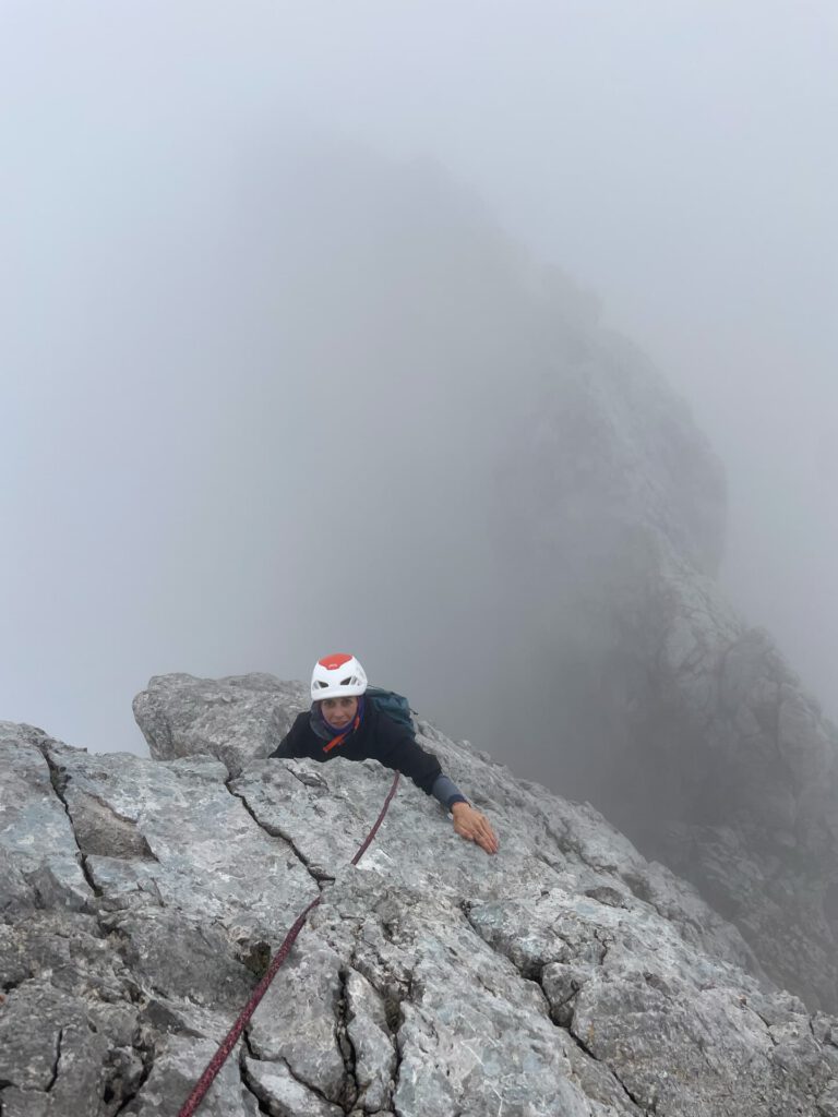 Eine Person mit Helm klettert am Hohen Dachstein auf dem Normalweg an einem Seil entlang eine steile, felsige Bergwand hinauf, während Nebel den Hintergrund und den Gipfel verdeckt.