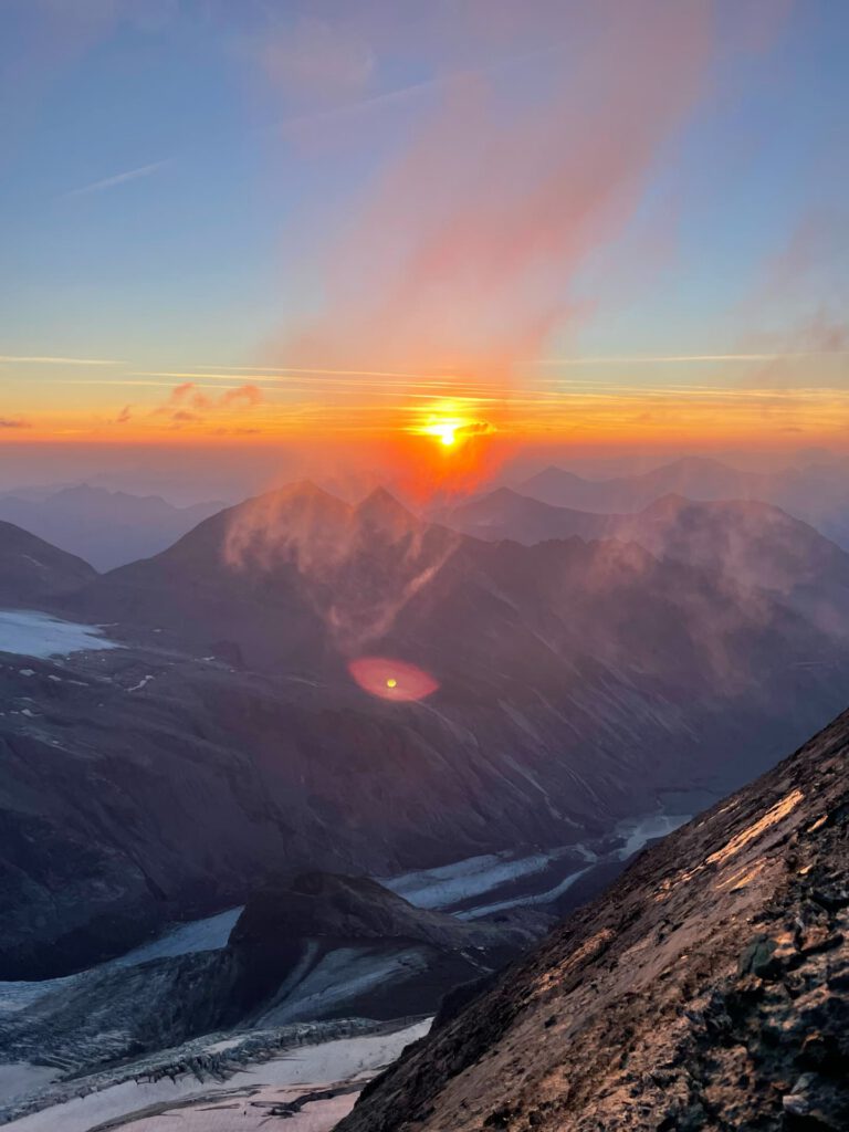 Die Sonne versinkt hinter den Berggipfeln und taucht den Himmel und die Wolken in ein orange-rosa Licht, während im Tal darunter Gletscher zu sehen sind – ein atemberaubender Anblick, der an ein „Glocknerwand geführt“-Abenteuer erinnert.