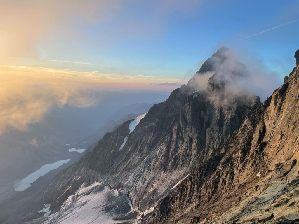 Ein steiler, felsiger Berggipfel mit Schneeflecken, der bei Sonnenaufgang oder Sonnenuntergang in goldenes Sonnenlicht getaucht ist. Wolken ziehen nahe dem Gipfel vorbei, von wo aus man auf ferne Seen und die nebelverhangenen Täler darunter blickt – eine Szenerie, die an ein Abenteuer auf der Glocknerwand erinnert.