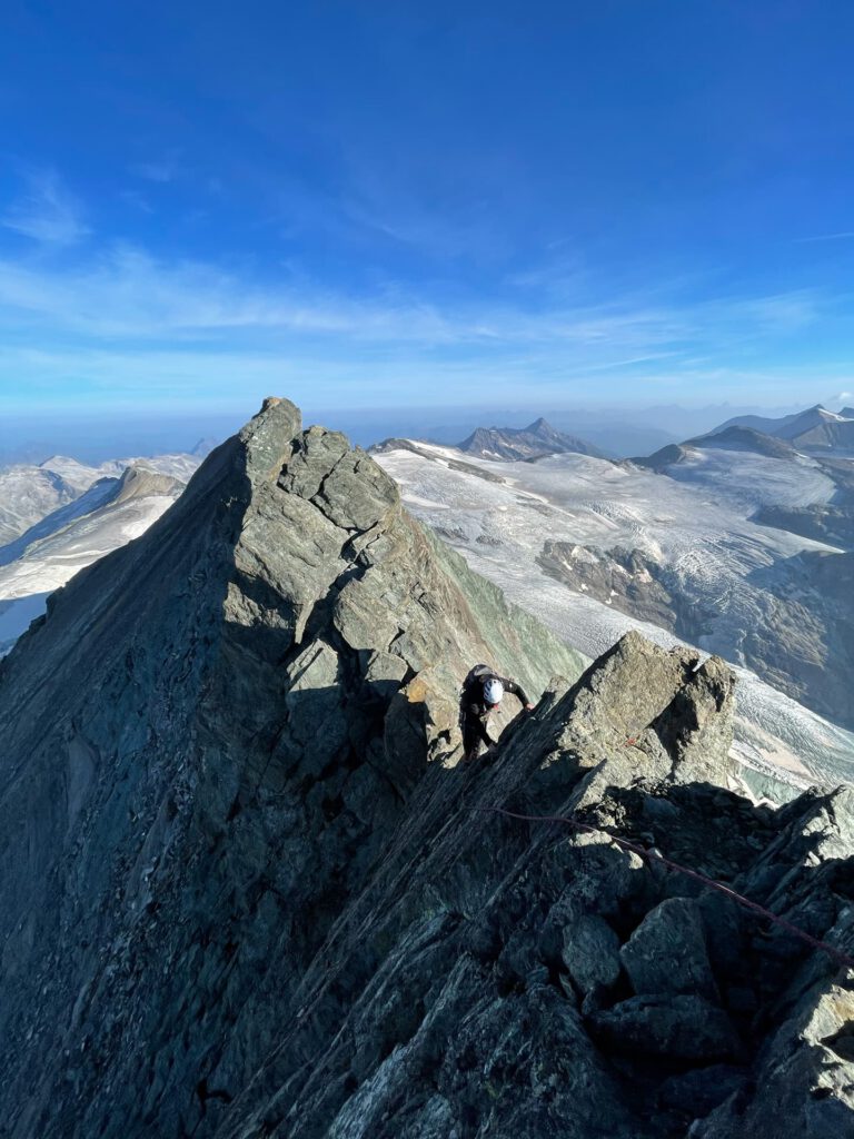 Ein Bergsteiger erklimmt auf einer geführten Tour an der Glocknerwand einen zerklüfteten, felsigen Berggrat unter strahlend blauem Himmel, umgeben von schneebedeckten Gipfeln und fernen Tälern.