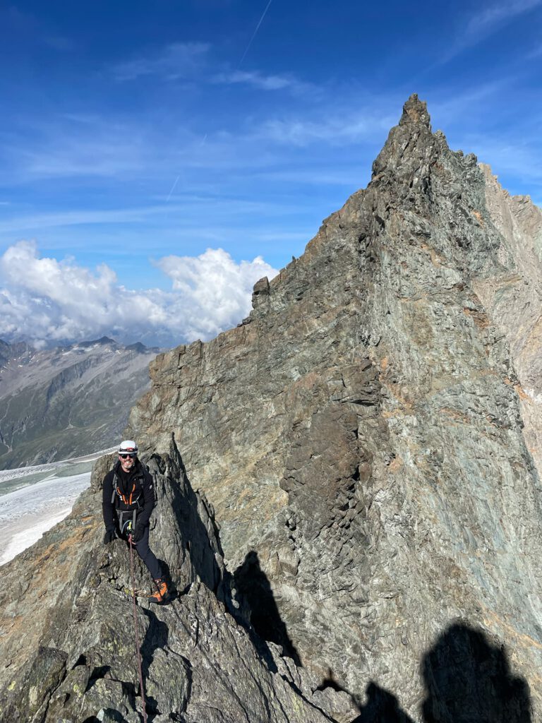 Ein Kletterer in Kletterausrüstung steht auf einem zerklüfteten, felsigen Berggrat unter einem blauen Himmel mit Wolken. Im Hintergrund sind schneebedeckte Gipfel und ein Gletscher zu sehen, die die dramatische Kulisse einer geführten Besteigung der Glocknerwand heraufbeschwören. Schatten fallen auf die Felsen.