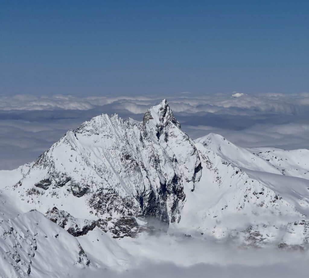 Ein schneebedeckter Berggipfel ragt steil unter einem strahlend blauen Himmel empor, umgeben von Wolkenschichten und fernen schneebedeckten Bergkämmen – eine atemberaubende Kulisse, die oft im Rahmen von geführten Bergtouren erkundet wird, bei denen erfahrene Bergführer Abenteurer durch diese unberührte Alpenlandschaft führen.