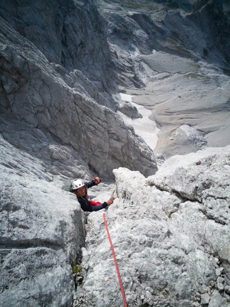 Zwei Kletterer, die Helme und Kletterausrüstung tragen, erklimmen eine steile, felsige Bergwand auf der berühmten Route „Steinerweg Dachstein geführt“. Ein rotes Seil verbindet sie, während sie sich ihren Weg nach oben bahnen, unter ihnen erstreckt sich eine dramatische, raue Landschaft.