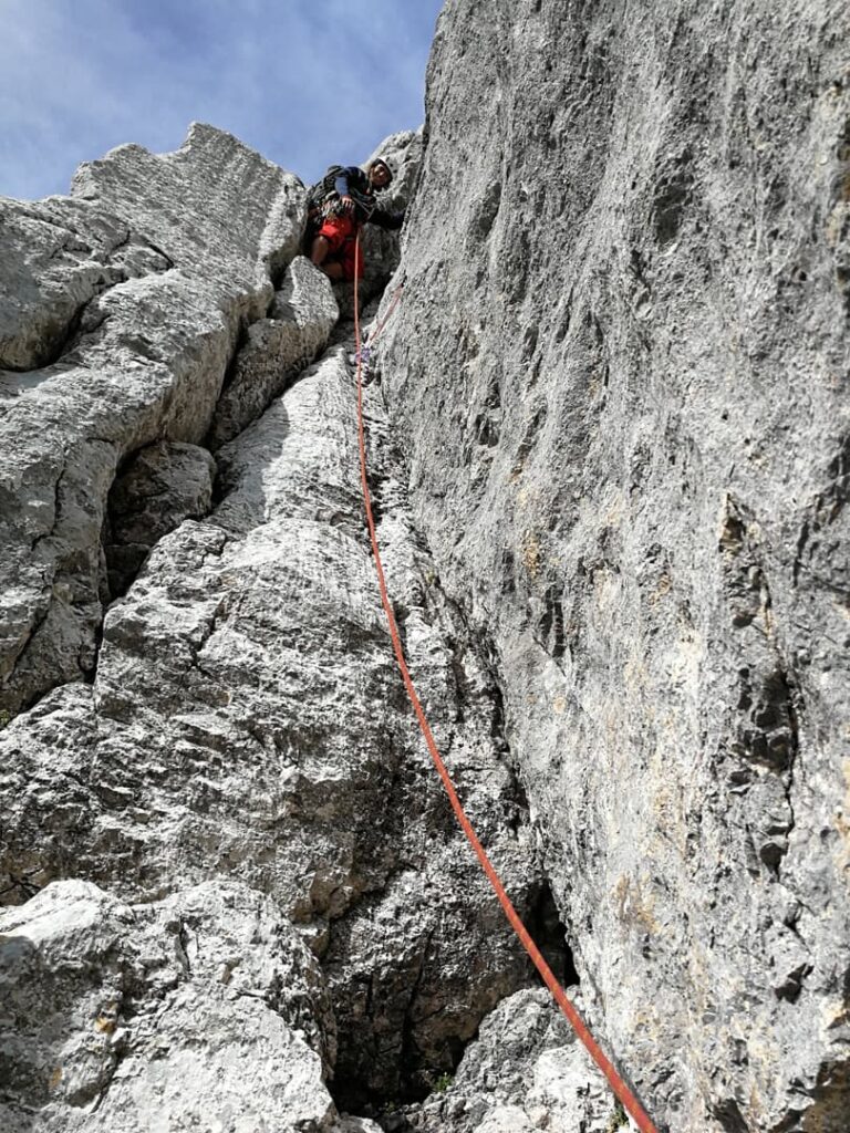 Eine Person in Kletterausrüstung erklimmt eine steile Felswand auf dem berühmten Steinerweg-Dachstein-Führungsweg, gesichert durch ein rotes Seil. Der Himmel ist blau mit vereinzelten Wolken, und schroffe graue Felsen prägen das Bild.