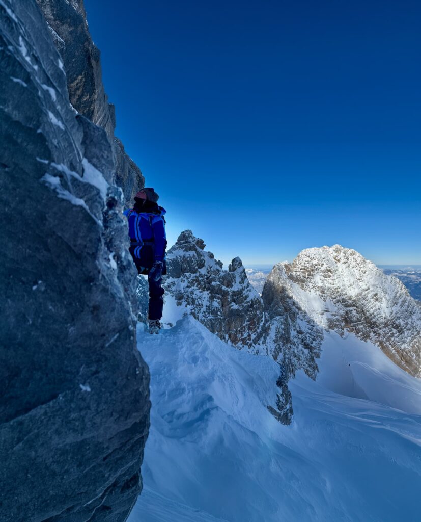 Ein Bergsteiger in Winterausrüstung steht auf einem verschneiten, schmalen Felsvorsprung entlang des Hoher-Dachstein-Normalwegs, im Hintergrund sind schroffe, schneebedeckte Gipfel und ein tiefblauer Himmel zu sehen.