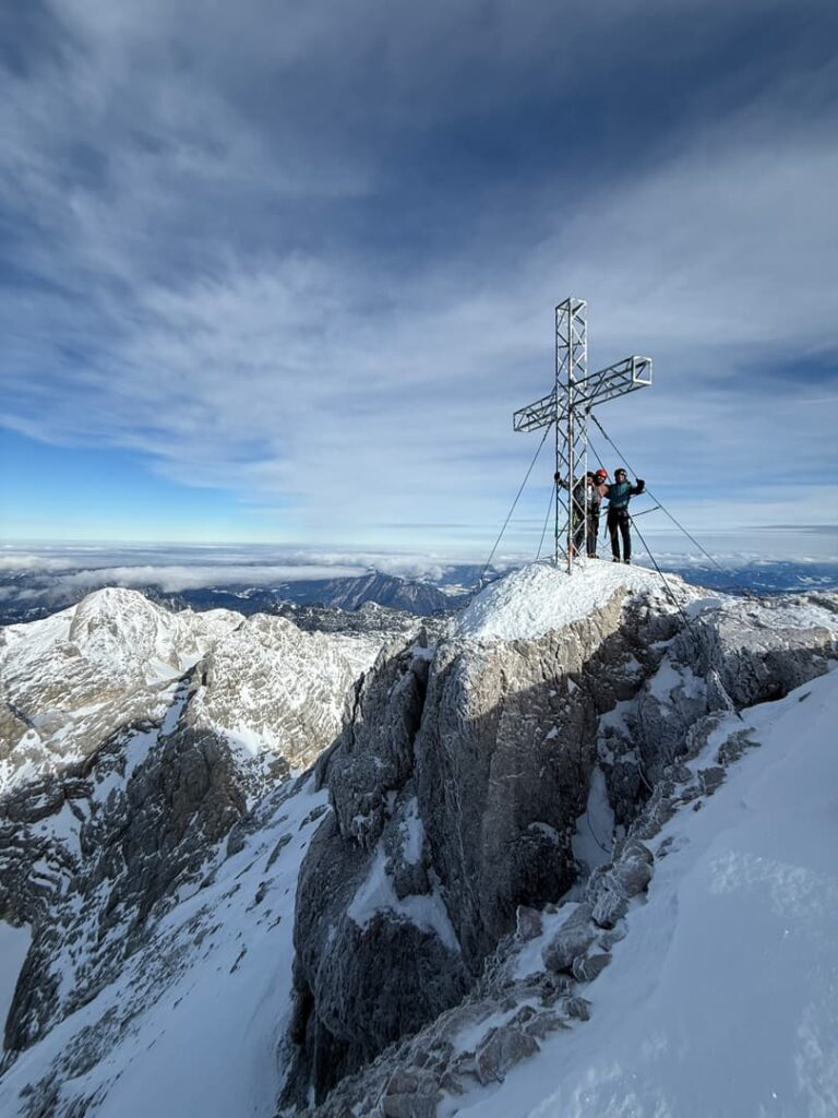 Zwei Menschen stehen neben einem großen Metallkreuz auf einem schneebedeckten Berggipfel, den sie über den Hoher-Dachstein-Normalweg erreicht haben, vor dem sich schroffe Grate und ein weiter, wolkenverhangener Himmel abzeichnen.