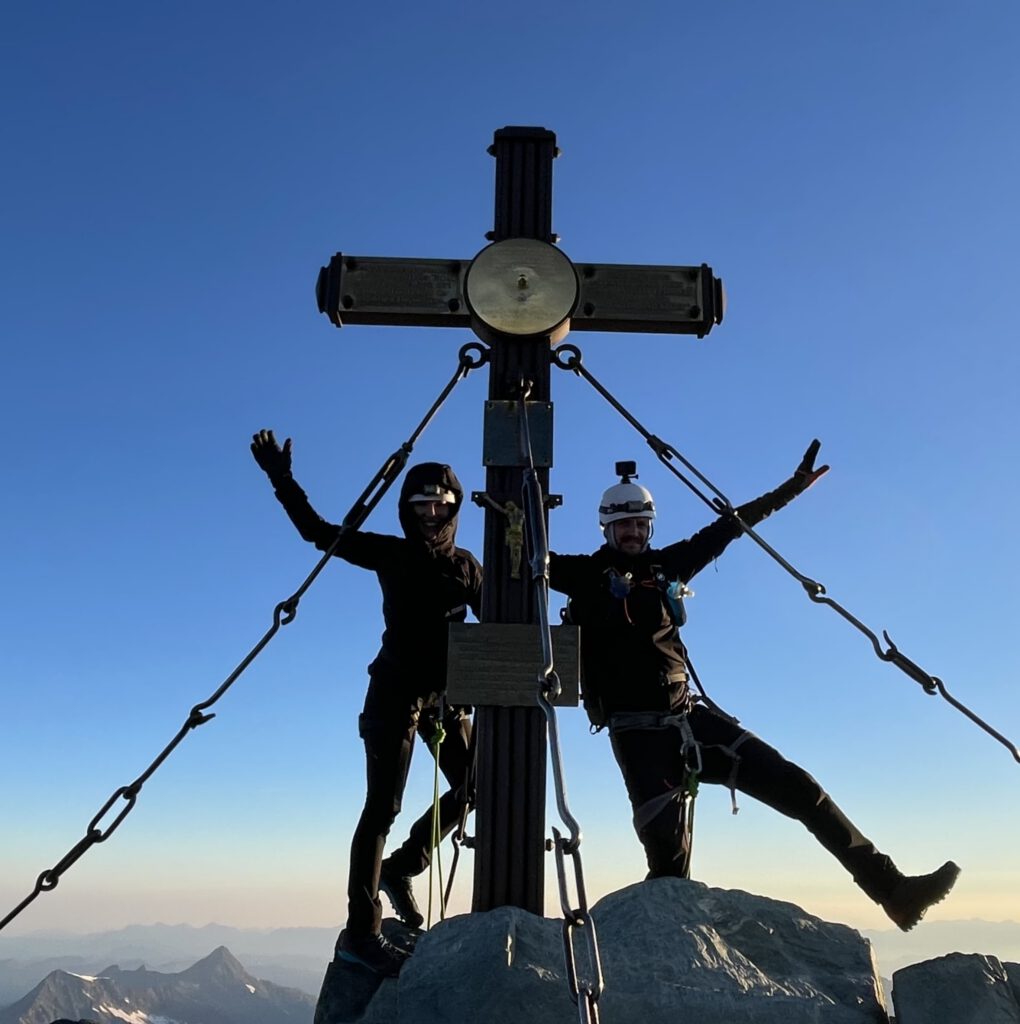 Zwei Bergsteiger mit Helmen und Ausrüstung posieren fröhlich auf einem Berggipfel und stehen neben einem großen Metallkreuz, das mit Ketten am Felsen verankert ist, vor einem strahlend blauen Himmel – ein Bild, das den Nervenkitzel und den Zusammenhalt einfängt, die geführte Bergtouren ausmachen.
