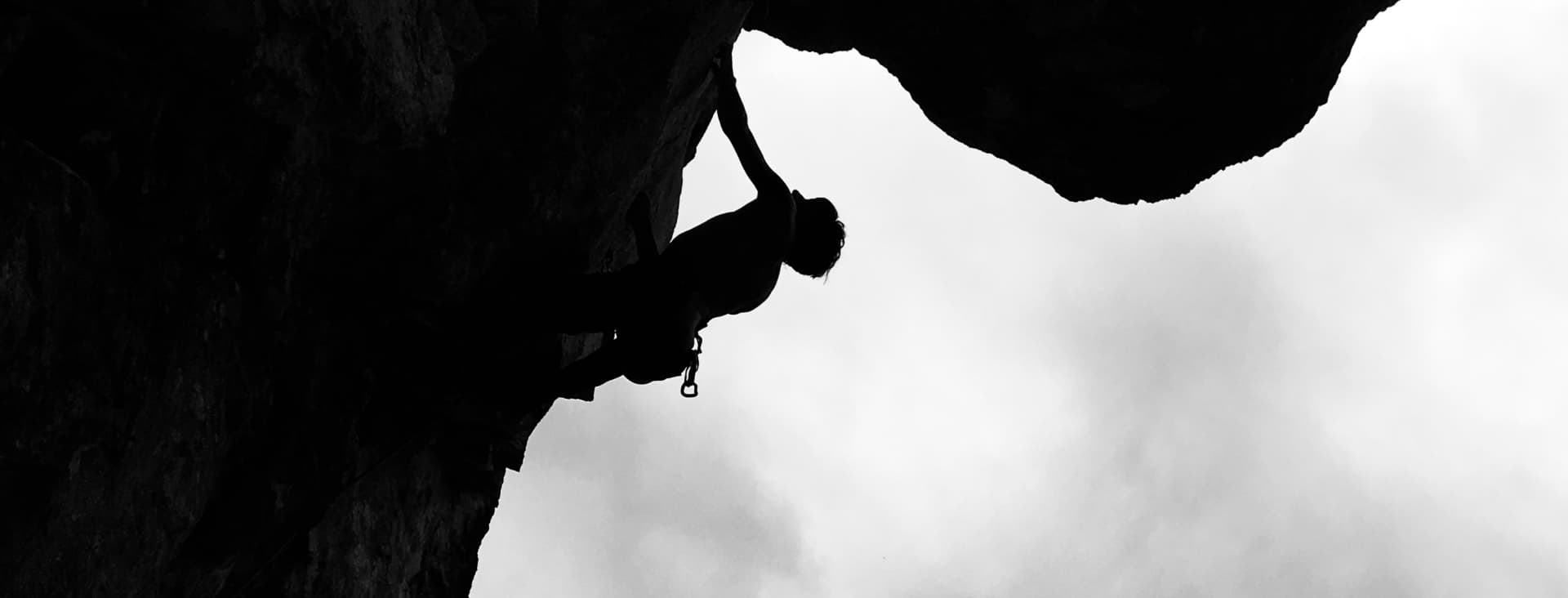 Die Silhouette eines Kletterers, der eine überhängende Felswand erklimmt, sich mit einem Arm festhält und sich vor einem bewölkten Himmel abzeichnet – die perfekte Inspiration für alle, die Fragen zu geführten Bergtouren haben.