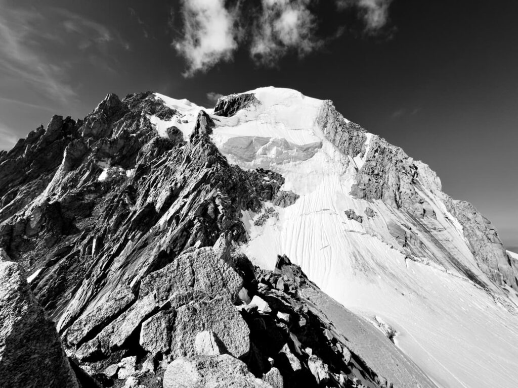 Schwarz-Weiß-Foto eines schroffen, schneebedeckten Berggipfels mit zerklüfteten Felsen im Vordergrund und einigen Wolken am Himmel, das die dramatischen Landschaften einfängt, die Thomas Fölsner Bergführer oft erkundet.