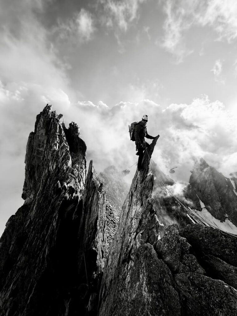 Ein Bergsteiger steht auf einem zerklüfteten Berggipfel, mit Helm und Rucksack, umgeben von dramatischen Wolken und schroffen Felsformationen. Dieses in eindrucksvollem Schwarz-Weiß aufgenommene Foto spiegelt den Abenteuergeist und die Fachkompetenz wider, für die Thomas Fölsner Bergführer steht.