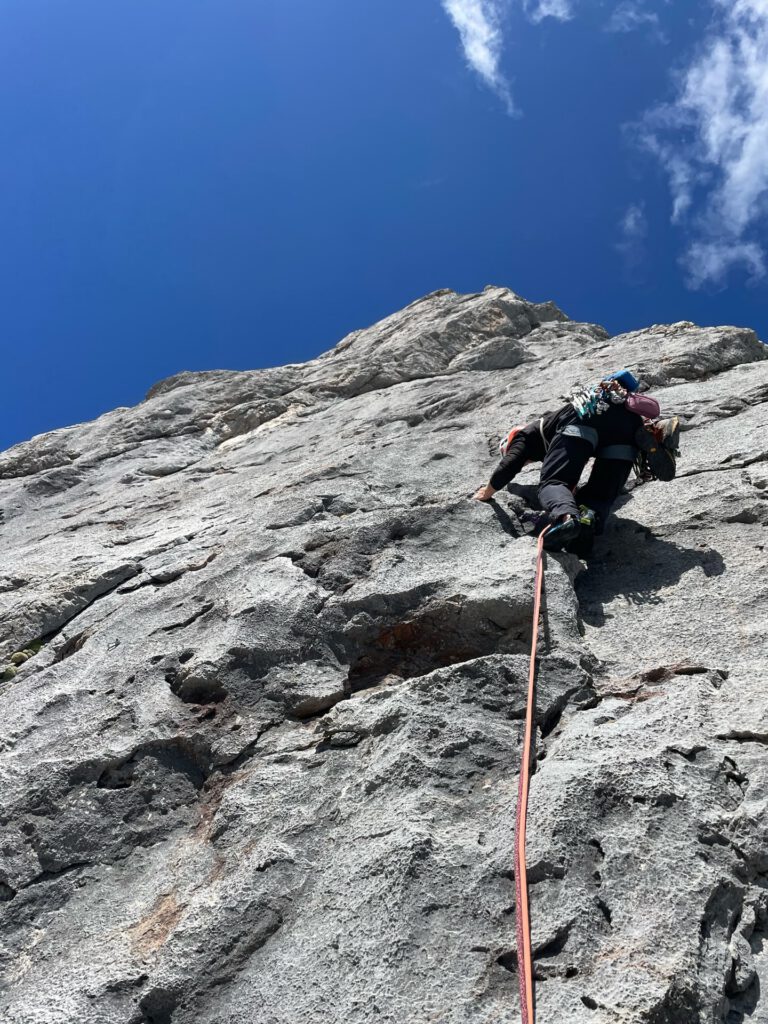 An einem sonnigen Tag erklimmt ein Kletterer eine steile, felsige Bergwand, gesichert mit Kletterausrüstung und einem roten Seil, das sich vor dem tiefblauen Himmel nach unten schlängelt – ein Aufstieg, der an die anspruchsvollen „Königsjodler“-Führungen erinnert.