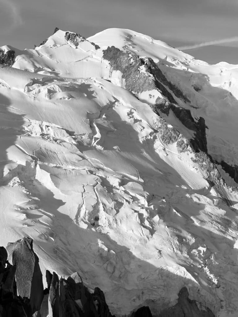 Ein schneebedeckter Berg mit zerklüfteten Gipfeln und Graten, teilweise im Schatten und teilweise im Sonnenlicht, unter einem bewölkten Himmel. Einige freiliegende Felsabschnitte durchbrechen die weiße Schneedecke und das Eis und erinnern an die dramatischen Ausblicke, die man auf einer geführten Steinerweg-Dachstein-Tour erleben kann.