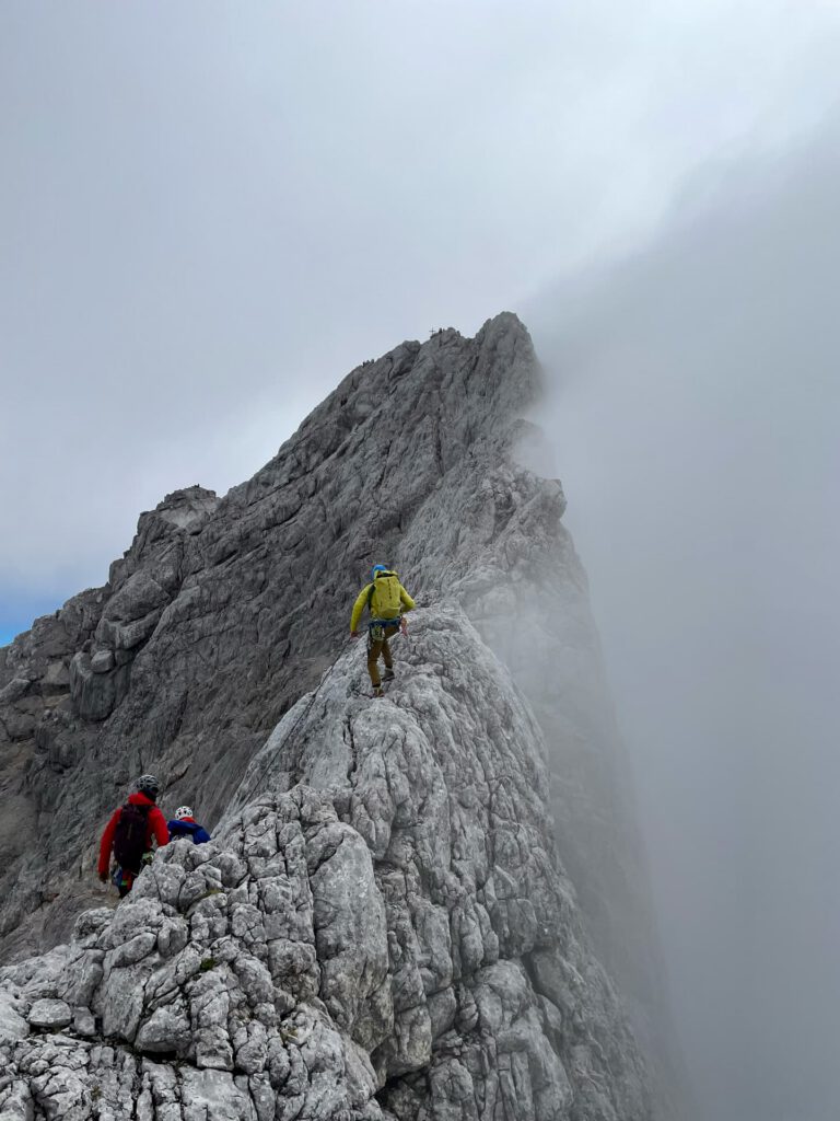Drei Bergsteiger in leuchtend bunten Jacken steigen auf dem Normalweg zum Hohen Dachstein einen schmalen, felsigen Berggrat hinauf, umgeben von Wolken und Nebel, mit steilen Abgründen zu beiden Seiten.