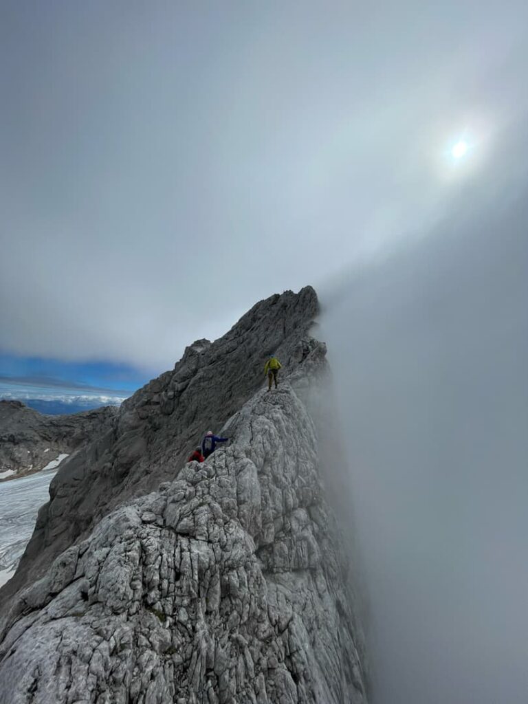 Zwei Bergsteiger in leuchtend bunten Jacken erklimmen einen zerklüfteten, felsigen Bergrücken – wahrscheinlich entlang der berühmten Route „Steinerweg Dachstein geführt“ – unter bewölktem Himmel, wobei Nebel einen Teil der Szene verdeckt und Sonnenlicht schwach durch die Wolken scheint.