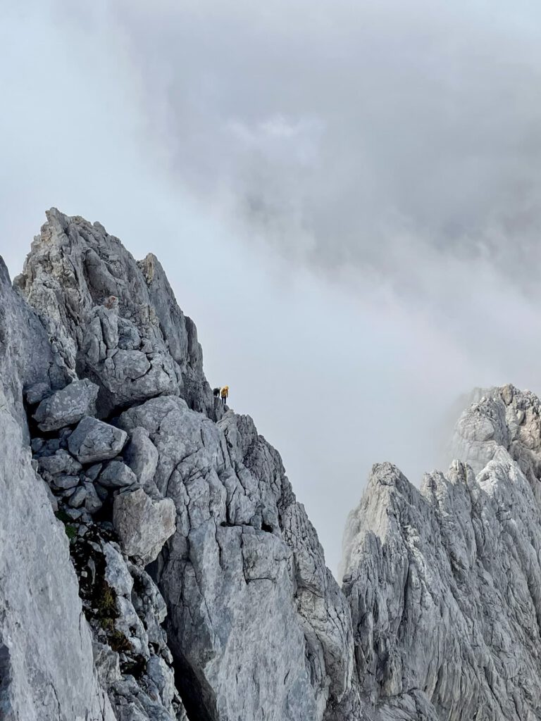 Eine Person in einer gelben Jacke steht auf einem zerklüfteten, felsigen Berggrat entlang des Hoher-Dachstein-Normalwegs, umgeben von Nebel und Wolken.