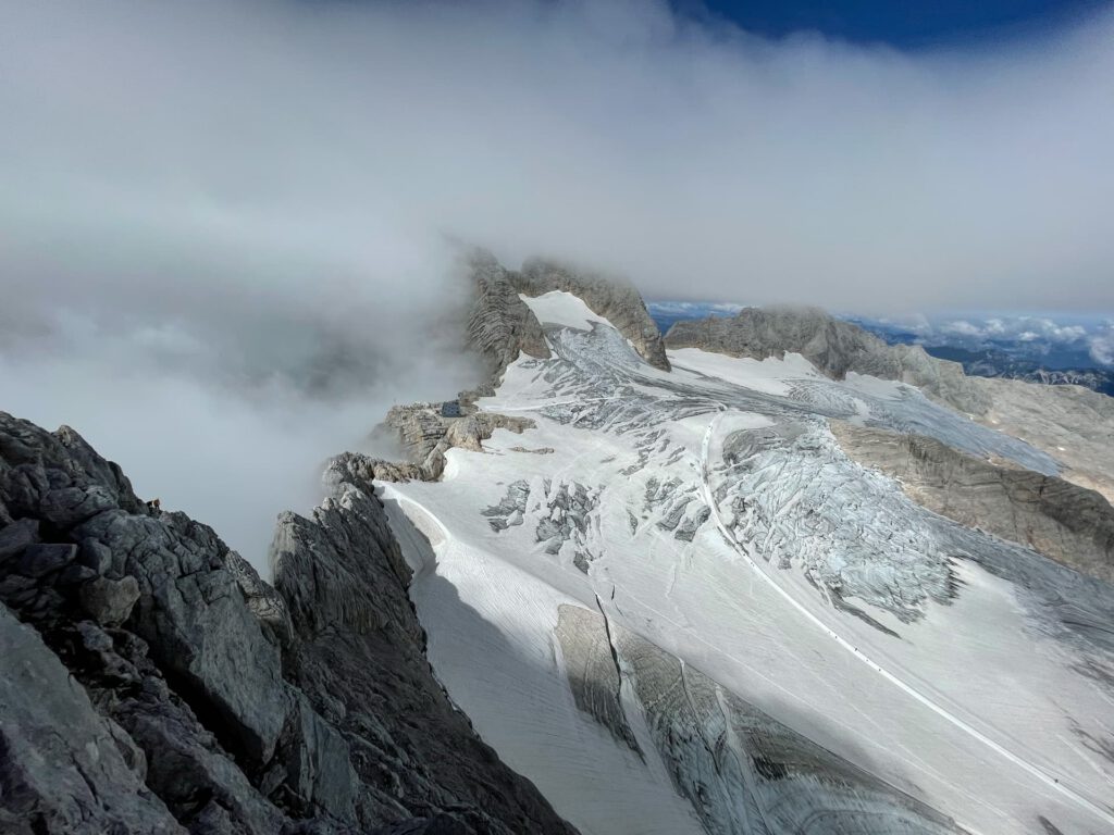 Schneebedeckte, teilweise in Wolken gehüllte Berggipfel ragen über schroffe Felshänge entlang des berühmten Hohen Dachstein-Normalwegs empor, während in der Ferne hier und da ein Stück blauer Himmel zu sehen ist.