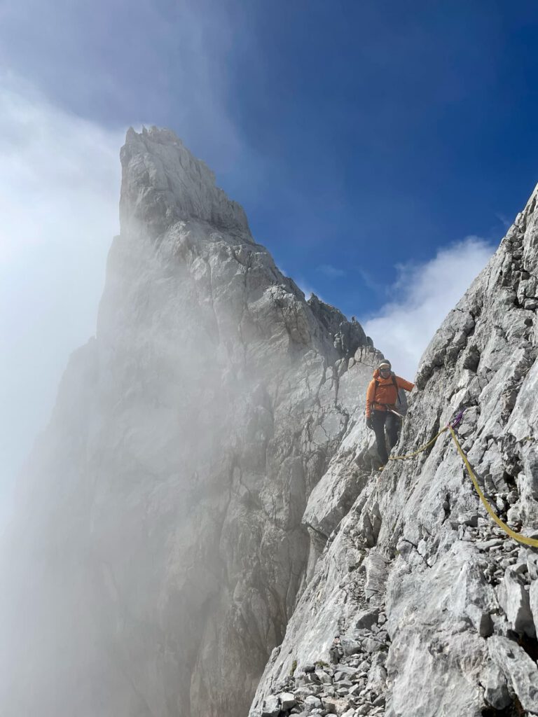 Ein Kletterer in einer orangefarbenen Jacke erklimmt unter blauem Himmel einen steilen, felsigen Berggrat entlang des Hohen Dachstein-Normalwegs, wobei Nebel und Wolken den Gipfel umhüllen und er sich zur Sicherheit an einem gesicherten Seil festhält.