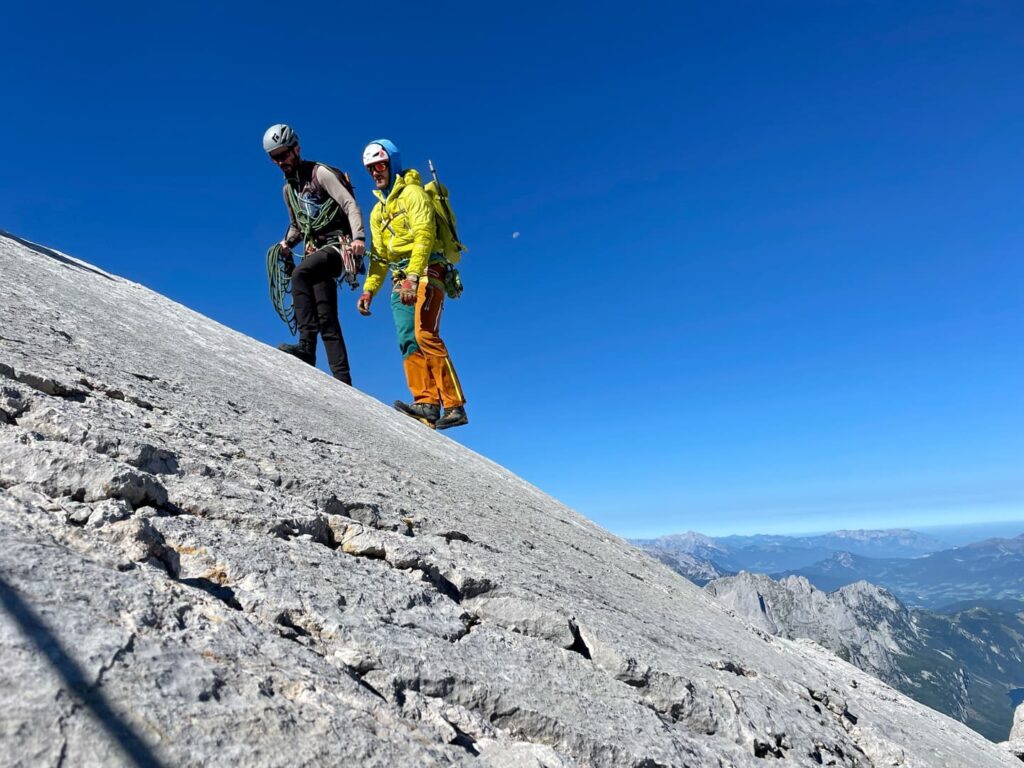 Zwei Bergsteiger mit Helmen und Ausrüstung stehen unter strahlend blauem Himmel auf einem steilen Felshang; sie haben gerade den Steinerweg am Dachstein hinter sich gebracht, und im Hintergrund sind Berge und Täler zu sehen.