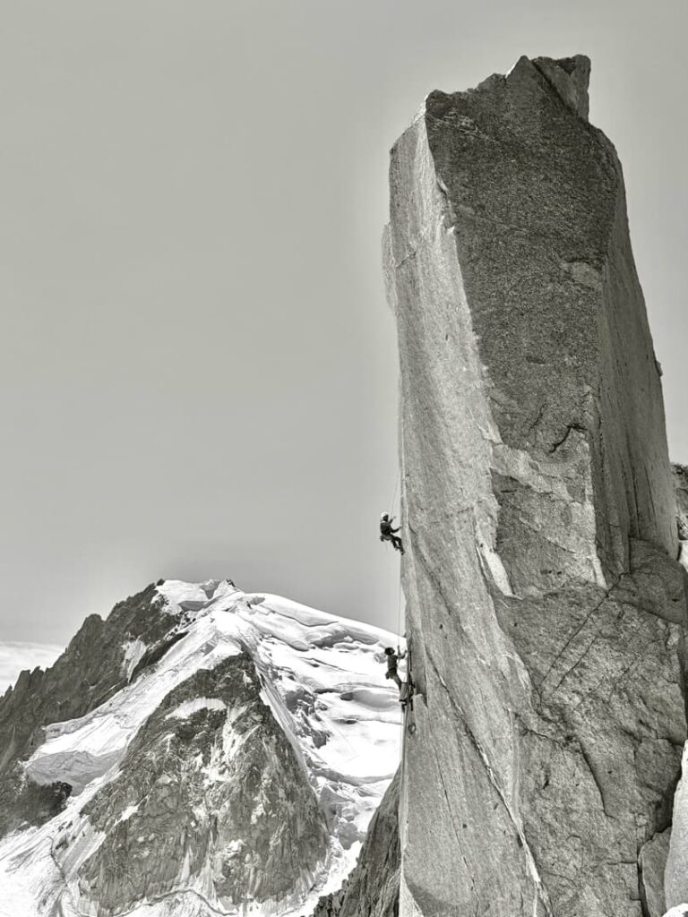 Zwei Kletterer erklimmen einen hohen, steilen Felsspitz entlang der berühmten Route „Steinerweg Dachstein geführt“, im Hintergrund sind schneebedeckte Berge vor klarem Himmel zu sehen.
