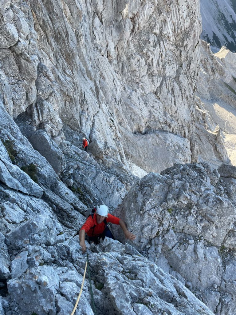 Zwei Kletterer mit Helmen und roten Hemden erklimmen auf dem Kletterweg „Pichlweg Dachstein geführt“ eine steile, felsige Bergwand, gesichert durch ein Seil. Unter dem klaren Himmel erhebt sich das zerklüftete Gelände steil um sie herum.