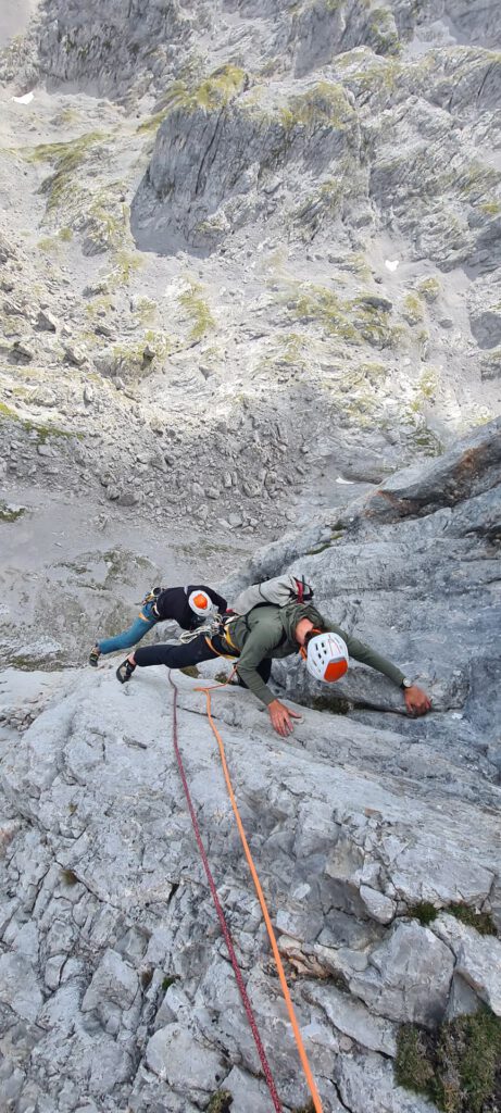 Zwei Bergsteiger mit Helmen und Klettergurten erklimmen auf der berühmten Route „Königsjodler geführt“ eine steile Felswand, gesichert durch Seile, während sich unter ihnen eine raue, graue Landschaft erstreckt.