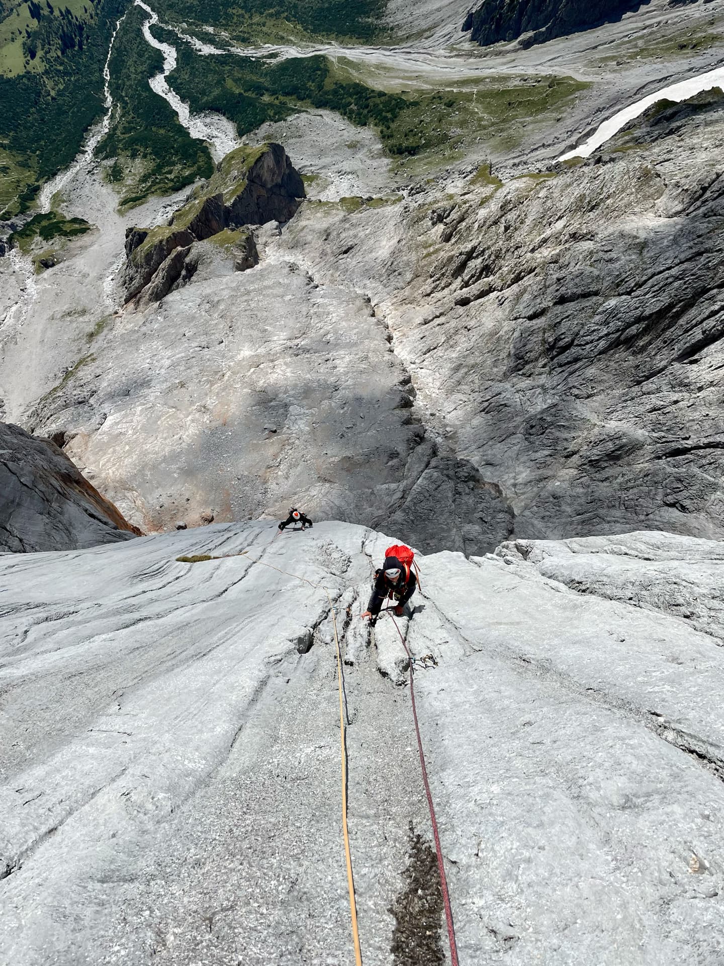 Zwei Kletterer erklimmen mit Seilen eine steile, graue Felswand auf der berühmten Route „Königsjodler geführt“, hoch über einer Berglandschaft mit grünen Wäldern und einem sich schlängelnden Fluss im Hintergrund.