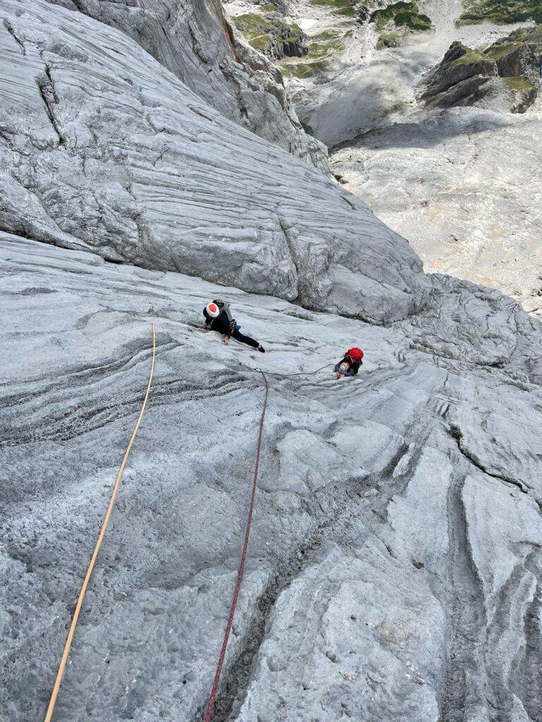 Zwei Kletterer mit Helmen erklimmen auf der berühmten Route „Königsjodler geführt“ mithilfe von Seilen eine steile, graue Felswand. Das Gelände wirkt schroff und strukturiert, unter ihnen sind dramatische Klippen und eine felsige Landschaft zu sehen.