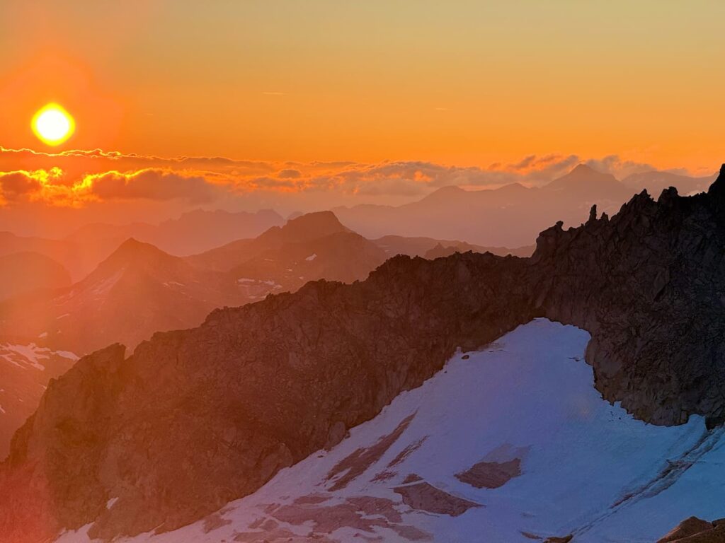 Sonnenuntergang über zerklüfteten Berggipfeln, wobei die Sonne teilweise von Wolken verdeckt wird. Schneeflecken bedecken Teile des dunklen Berghangs im Vordergrund und erinnern an Szenen aus einer Skitour auf den Großvenediger, während die fernen Bergketten im orange-rosa Himmel verblassen.