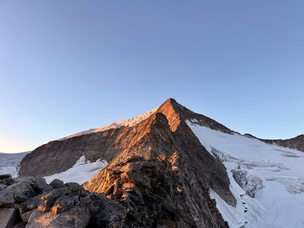 Ein felsiger Berggipfel liegt im warmen Sonnenlicht, während Schnee und Eis unter einem strahlend blauen Himmel seine Hänge bedecken. Schroffe Felsen prägen den Vordergrund und unterstreichen die raue Alpenlandschaft – eine inspirierende Szene, die an ein Skitourenabenteuer auf dem Großvenediger erinnert.