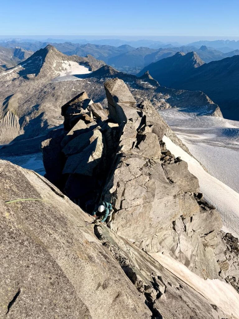 Ein Kletterer mit Helm erklimmt unter strahlend blauem Himmel einen steilen, felsigen Berggrat mit schroffen Gipfeln und einem Gletscher im Hintergrund und vermittelt so den Nervenkitzel eines Abenteuers auf der Skitour zum Großvenediger.