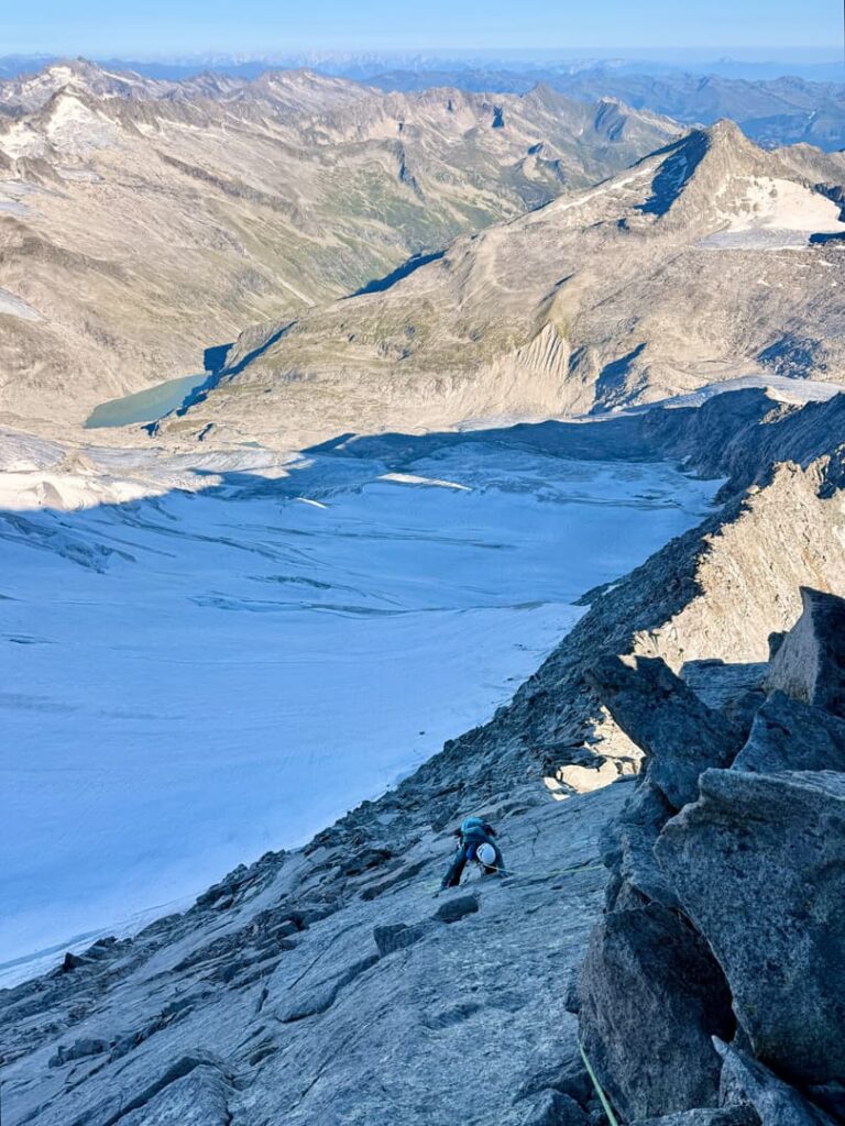 Ein Bergsteiger erklimmt einen steilen Felshang, unter ihm liegen Schnee und ein Gletscher; umgeben von schroffen Alpenbergen und fernen Tälern unter strahlend blauem Himmel – eine Szene, die an eine Skitour auf den Großvenediger erinnert.