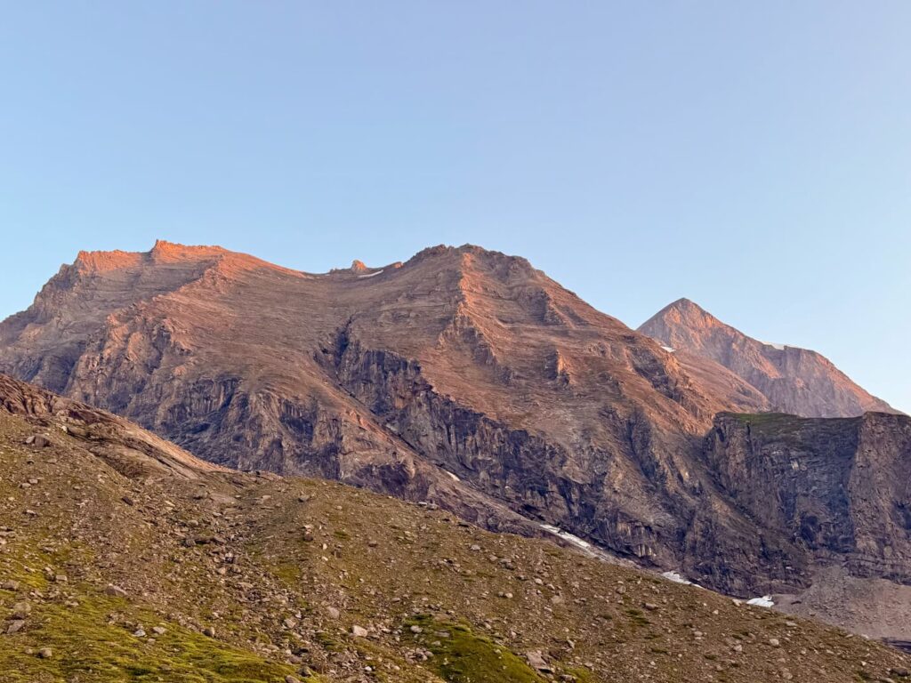 Das Sonnenlicht taucht die schroffen Berggipfel des Großen Wiesbachhorns unter einem strahlend blauen Himmel in einen warmen Schein, während im Vordergrund felsiges Gelände und grüne Vegetationsflecken zu sehen sind.